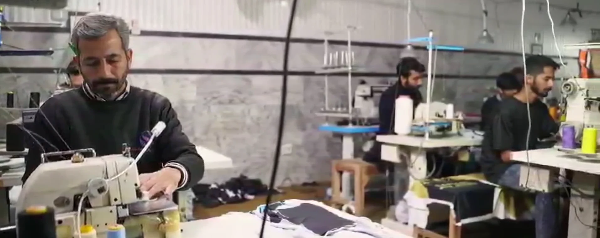 Drain Sport production floor in Sialkot: an operator at an overlock machine, the stitching line, sublimation printing, cutting, and garments being packed into cartons
